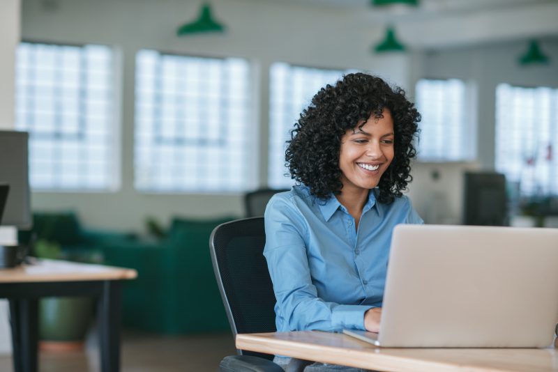 Smiling young businesswoman sitting at her desk in a large modern office working online with a laptop