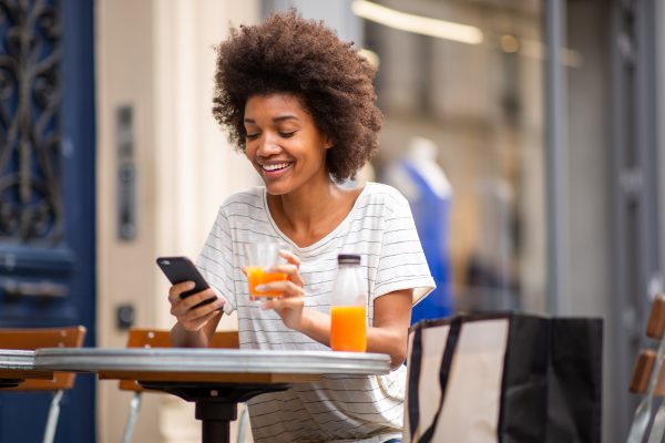 Portrait of beautiful young black woman sitting at outdoor cafe with cellphone and drink